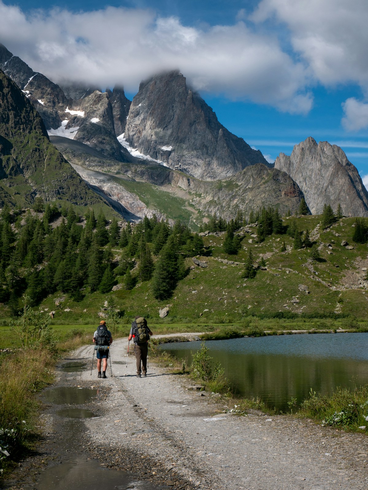 Two walkers following a snow-free mountain trail beside a lake