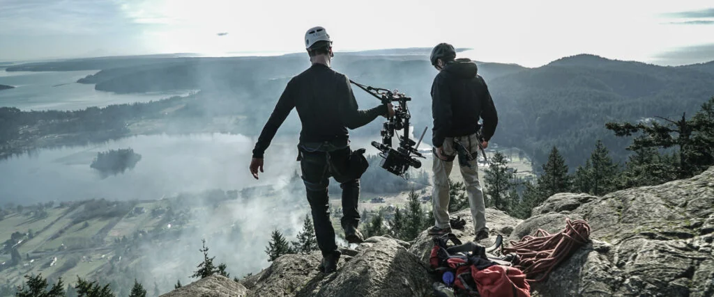 Helmeted film crew with camera equipment and ropes at a cliff-top location