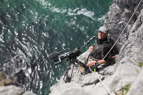 A camera operator supported on ropes beside a coastal cliff
